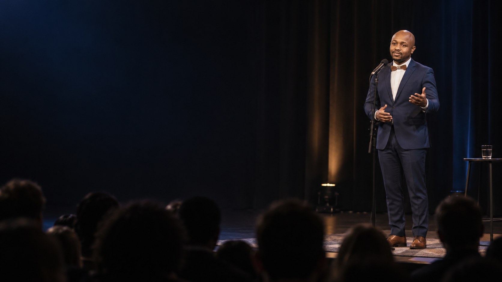 Ryan Perez speaking at a microphone stand on a dark stage, audience in silhouette